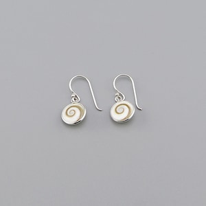 Puede incluir: Un par de pendientes colgantes de plata. Cada pendiente presenta un colgante circular blanco con un diseño en espiral crema. Los pendientes tienen un cierre de gancho simple y se muestran sobre un fondo gris.