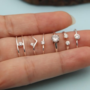 May include: A collection of silver rings with various designs, including a solitaire, a chevron, and rings with small, round gemstones. The rings are displayed on a hand against a light blue background. The rings are delicate and minimalist.