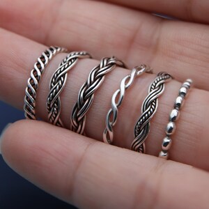 Könnte beinhalten: Eine Sammlung von Silberringen mit aufwendigen geflochtenen und strukturierten Designs. Die Ringe weisen verschiedene Stile auf, darunter seilartige Drehungen, Webmuster und ein Perlenband. Die Ringe werden auf einem Finger präsentiert, um die verschiedenen Designs hervorzuheben.