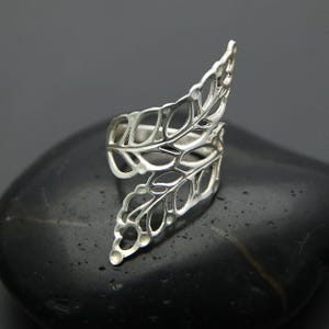 Könnte beinhalten: Ein silberfarbener Ring mit Blattmuster. Der Ring hat ein durchbrochenes Design mit mehreren Blattformen, wodurch eine zarte und von der Natur inspirierte Ästhetik entsteht. Der Ring ist vor einem dunklen Hintergrund platziert.
