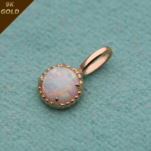 Può includere: Ciondolo in oro con un'opale rotonda e iridescente. La pietra è incastonata in una lunetta smerlata e ha un'asola per una catena. Il testo "9K GOLD" è visibile in alto a sinistra.