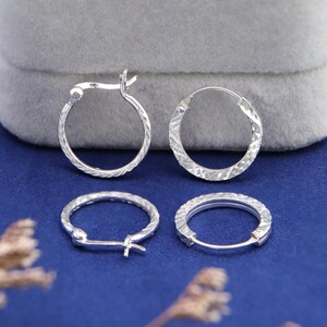 May include: Four silver hoop earrings with a textured, diamond-cut pattern. Two earrings are open, and two are closed, showing the clasp. The earrings are on a blue surface with a grey background.