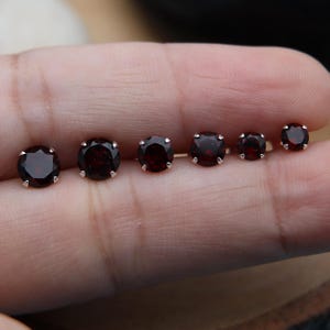 May include: A collection of six round garnet stud earrings in varying sizes. Each earring features a deep red gemstone set in a silver-colored metal. The earrings are displayed in a row, showcasing the size differences.