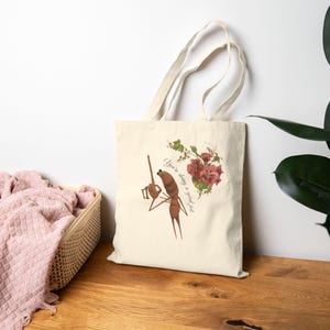 May include: Cream-colored tote bag featuring a brown ant-like figure holding a microphone and sitting on a chair. The bag has a floral design with red flowers and the text "You're doing a good job."