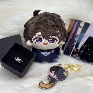 May include: An assortment of merchandise including a plush doll with brown hair and a blue suit, a silver ring in a black box, a keychain with a cartoon character, and several photo cards. The items are arranged on a white, fluffy surface.