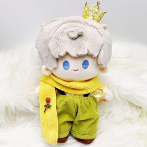 Puede incluir: Muñeco de peluche de un personaje con cabello gris claro, ojos azules y una pequeña sonrisa. El muñeco lleva una corona dorada, una bufanda amarilla con una rosa, una camisa amarilla y pantalones de pana verdes. El muñeco está sobre una superficie blanca y esponjosa.