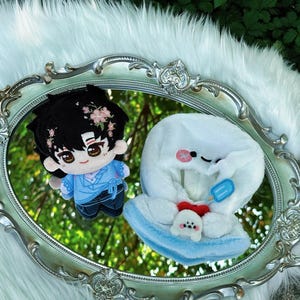 May include: Two plush dolls are reflected in a silver-framed mirror. One doll is a boy with black hair and pink flower accents, wearing a blue outfit. The other is a white plush with a blue popsicle and a small dog.