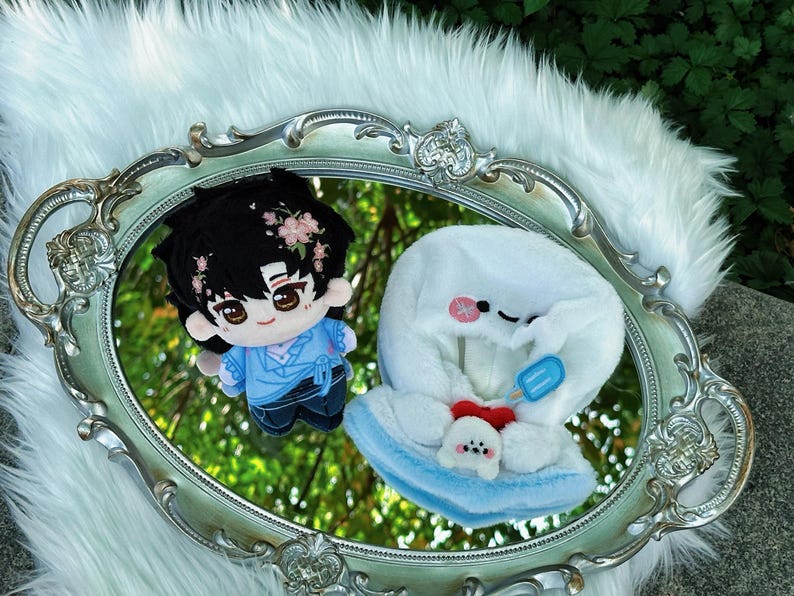 May include: Two plush dolls are reflected in a silver-framed mirror. One doll is a boy with black hair and pink flower accents, wearing a blue outfit. The other is a white plush with a blue popsicle and a small dog.