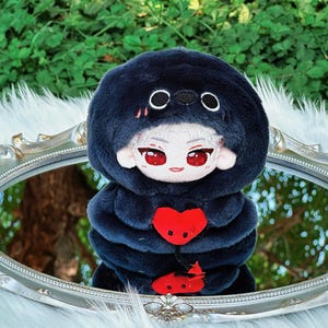May include: A plush doll with a dark blue hooded outfit and a red heart-shaped design. The doll has a white face with red eyes and is reflected in a silver-framed mirror. The background features green foliage.