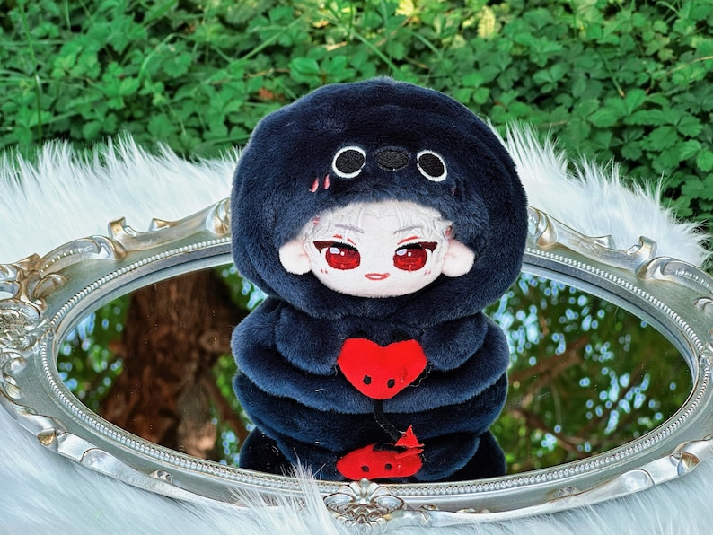 May include: A plush doll with a dark blue hooded outfit and a red heart-shaped design. The doll has a white face with red eyes and is reflected in a silver-framed mirror. The background features green foliage.