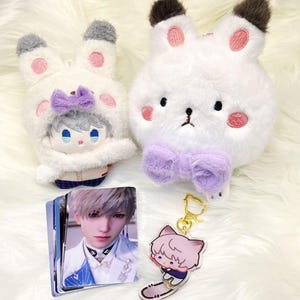 以下が含まれることがあります： ぬいぐるみとアクセサリーのコレクション。 ピンクのアクセントと紫のリボンが付いた2つの白いウサギ型のぬいぐるみ、キャラクターカードの山、猫をテーマにしたキーホルダーが含まれています。 ぬいぐるみは黒い目と茶色の耳の先端を持っています。