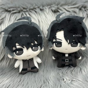 Puede incluir: Dos muñecos de peluche con pelo negro y gris y ojos grandes. Uno de los muñecos lleva un abrigo blanco sobre un conjunto gris, el otro lleva un conjunto negro. Los muñecos están sobre una superficie gris y peluda.