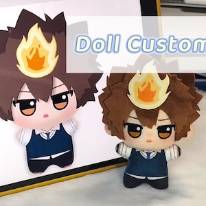 以下が含まれることがあります： 茶色の髪と炎の形をしたアクセサリーが付いたカスタムドール。人形は白いシャツの上に青いベストを着ており、大きな目があります。「Doll Custom」というテキストが人形の上に表示されています。