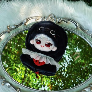 May include: A plush doll with a black hooded outfit and a white ruffled collar. The doll has a white face with red eyes and a red heart design on the front. It is set on a decorative oval mirror with a white fur background.