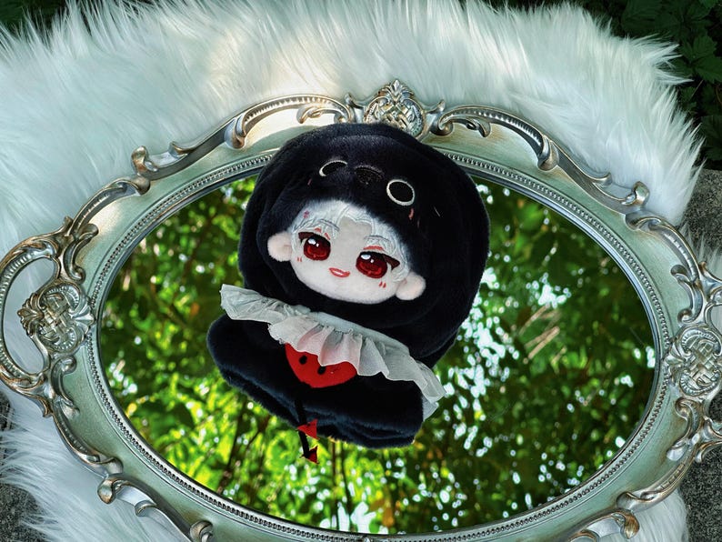 May include: A plush doll with a black hooded outfit and a white ruffled collar. The doll has a white face with red eyes and a red heart design on the front. It is set on a decorative oval mirror with a white fur background.