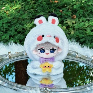 May include: A plush doll with a white bunny-eared hood, red cheeks, and a purple bow tie. The doll is holding a yellow star-shaped plush. The doll is reflected in a silver-framed mirror.