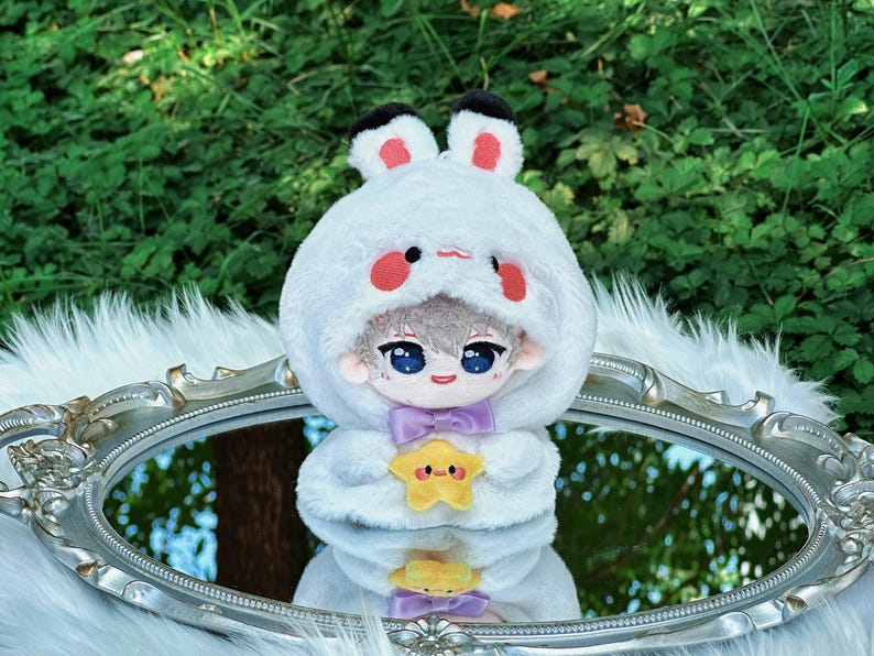 May include: A plush doll with a white bunny-eared hood, red cheeks, and a purple bow tie. The doll is holding a yellow star-shaped plush. The doll is reflected in a silver-framed mirror.