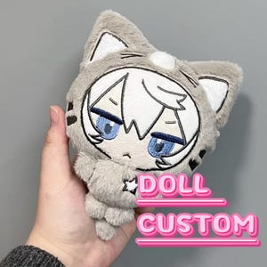 May include: A plush doll with a cat-eared hood, featuring an anime-style face with blue eyes and white hair. The doll is gray with black and white accents. The text "DOLL CUSTOM" is displayed in pink.