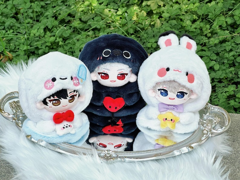 May include: Three plush dolls with anime-style faces, each wearing a hooded outfit. The dolls are on a silver tray with a white fur-like surface. The outfits are white, navy blue, and white with bunny ears. The dolls have red, blue, and brown eyes.