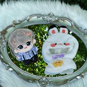 May include: Two plush dolls rest on a silver, ornate mirror. One doll is a boy with gray hair and floral accents, wearing a blue shirt and black pants. The other is a white bunny-shaped hood with a purple bow and a yellow star.