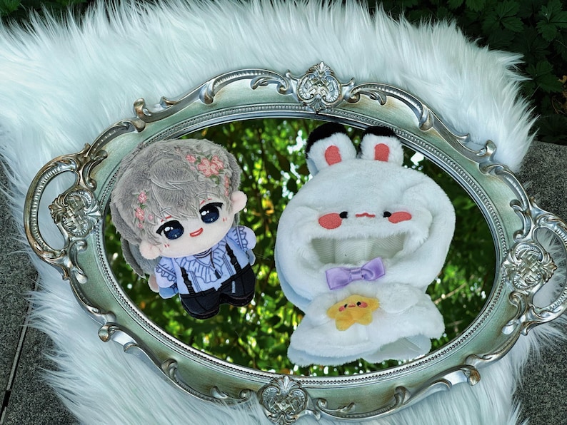 May include: Two plush dolls rest on a silver, ornate mirror. One doll is a boy with gray hair and floral accents, wearing a blue shirt and black pants. The other is a white bunny-shaped hood with a purple bow and a yellow star.