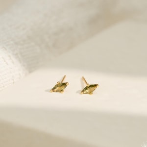 May include: A pair of gold-colored stud earrings with olive-green marquise-shaped stones. The earrings are set on a white surface with a soft, neutral background. The Sunny Day Jewelry logo is visible in the bottom right corner.