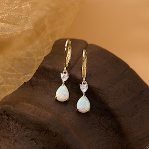 May include: A pair of gold-colored drop earrings. Each earring features a pear-shaped opal stone suspended below a heart-shaped clear stone. The earrings are displayed on a dark brown wooden surface with a gold-colored fabric in the background.