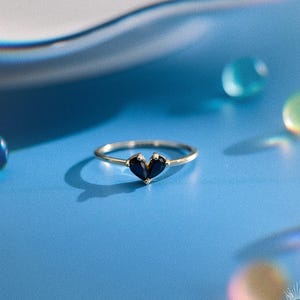 May include: A thin gold ring featuring two small, dark blue, pear-shaped gemstones set together to form a heart shape. The ring is centered on a smooth blue surface, surrounded by several out-of-focus, colorful, translucent circular spheres.