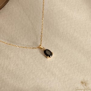 May include: A gold-coloured necklace featuring a small, black oval pendant. The delicate chain supports the pendant, which is set in a gold bezel. The necklace is presented on a neutral-toned surface. The Sunny Day logo is in the bottom right corner.