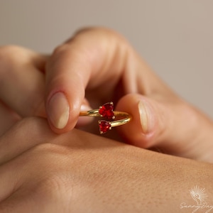 May include: A gold-colored ring featuring two heart-shaped, red gemstones. The ring's design curves around the finger, with the gemstones positioned at the top. The ring is held in a hand, showcasing the jewelry's design and color.