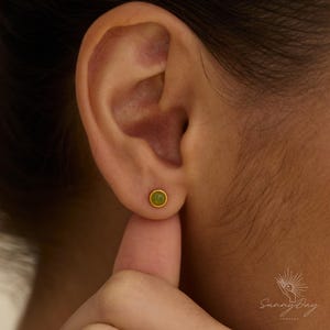 May include: A small, round stud earring featuring a smooth, olive-green cabochon stone set within a simple gold-toned metal bezel. The earring is positioned centrally on the earlobe, showcasing a minimalist design with a polished finish.