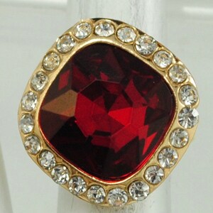 Peut inclure: Une bague dor&eacute;e orn&eacute;e d'une grosse pierre rouge fonc&eacute; taill&eacute;e en forme carr&eacute;e. La pierre est entour&eacute;e de petits cristaux ronds et transparents. La bague est pos&eacute;e sur un fond neutre, avec un design classique.