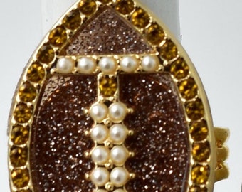 May include: Gold-toned ring with an oval-shaped face. The center features a sparkling brown inlay with a pearl and gold design. The ring is bordered by amber-colored gemstones, creating a glamorous accessory.