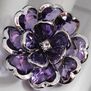 Könnte beinhalten: Eine silberne und violette Blumenbrosche mit einem Strassstein in der Mitte. Die Blume hat ein mehrschichtiges Design mit mehreren Blütenblättern.