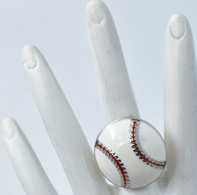 May include: A baseball-themed ring featuring a white enamel baseball design with red stitching and a silver-tone band. The ring is a unique accessory for sports fans.