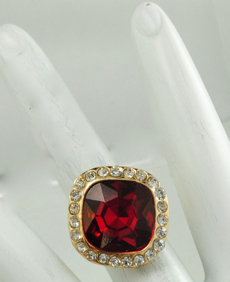 Peut inclure: Une bague dor&eacute;e orn&eacute;e d'une grosse pierre rouge fonc&eacute; carr&eacute;e et facett&eacute;e. La pierre est entour&eacute;e de petits cristaux transparents. La bague est pr&eacute;sent&eacute;e sur une surface blanche.