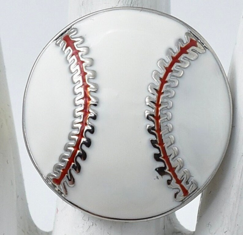 May include: A baseball-themed ring with a white enamel surface and red stitching detail. The ring has a silver-colored metal border. The design is a close-up of a baseball.
