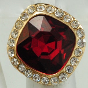 Peut inclure: Bague dor&eacute;e orn&eacute;e d'une grosse pierre rouge fonc&eacute; taill&eacute;e en carr&eacute;. La pierre est entour&eacute;e de petits cristaux clairs. Le sertissage de la bague est de couleur dor&eacute;e, cr&eacute;ant un contraste saisissant.