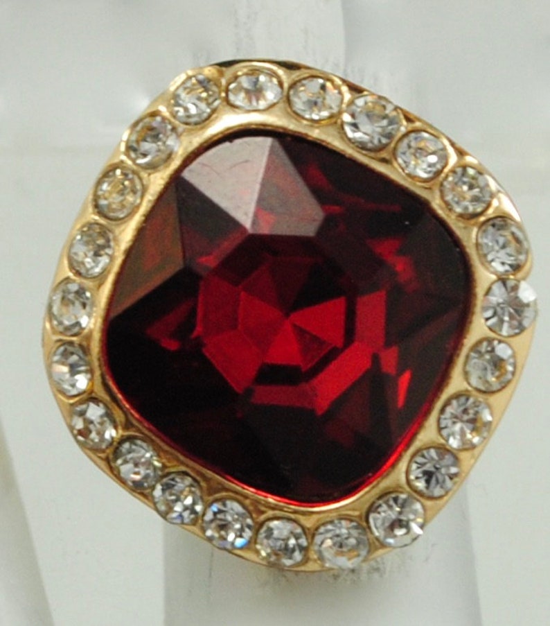 Peut inclure: Bague dor&eacute;e orn&eacute;e d'une grosse pierre rouge fonc&eacute; taill&eacute;e en carr&eacute;. La pierre est entour&eacute;e de petits cristaux clairs. Le sertissage de la bague est de couleur dor&eacute;e, cr&eacute;ant un contraste saisissant.