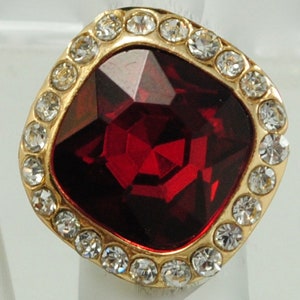 Peut inclure: Une bague dor&eacute;e orn&eacute;e d'une grosse pierre pr&eacute;cieuse rouge fonc&eacute; de forme carr&eacute;e au centre. La pierre pr&eacute;cieuse est entour&eacute;e d'une bordure de petits cristaux ronds et transparents. Le sertissage de la bague est dor&eacute;.