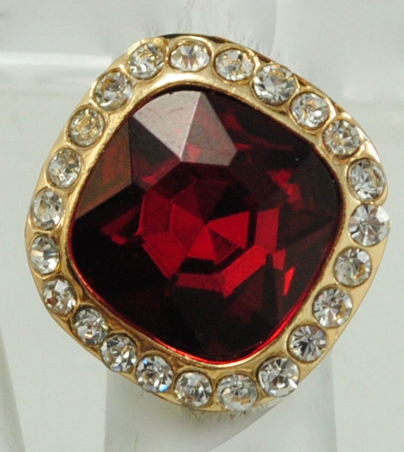 Peut inclure: Une bague dor&eacute;e orn&eacute;e d'une grosse pierre pr&eacute;cieuse rouge fonc&eacute; de forme carr&eacute;e au centre. La pierre pr&eacute;cieuse est entour&eacute;e d'une bordure de petits cristaux ronds et transparents. Le sertissage de la bague est dor&eacute;.