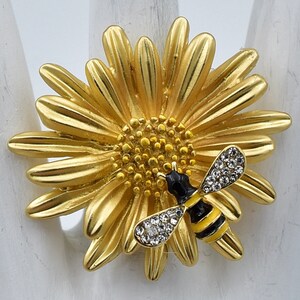 May include: A gold-toned ring featuring a large, textured daisy flower design. A small bee with black and yellow enamel stripes and clear crystal-encrusted wings is positioned on the petals of the flower, adding detail to the floral motif.