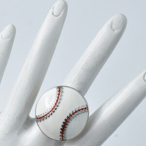 May include: A baseball-themed ring with a white enamel face and red stitching detail. The ring has a silver-colored metal band. The ring is round and sits on a white surface.