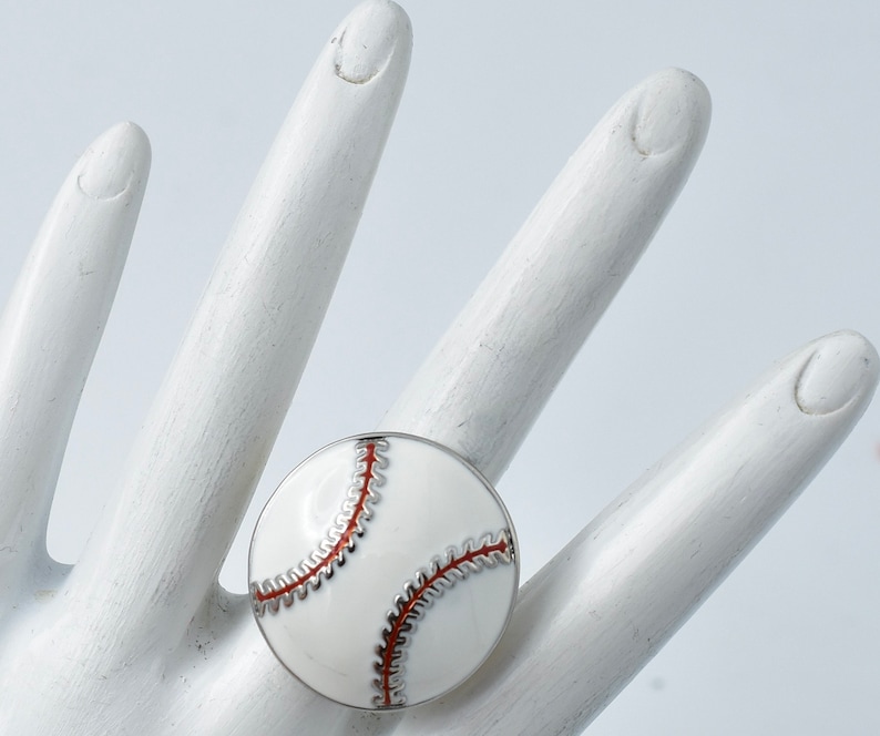 May include: A baseball-themed ring with a white enamel face and red stitching detail. The ring has a silver-colored metal band. The ring is round and sits on a white surface.