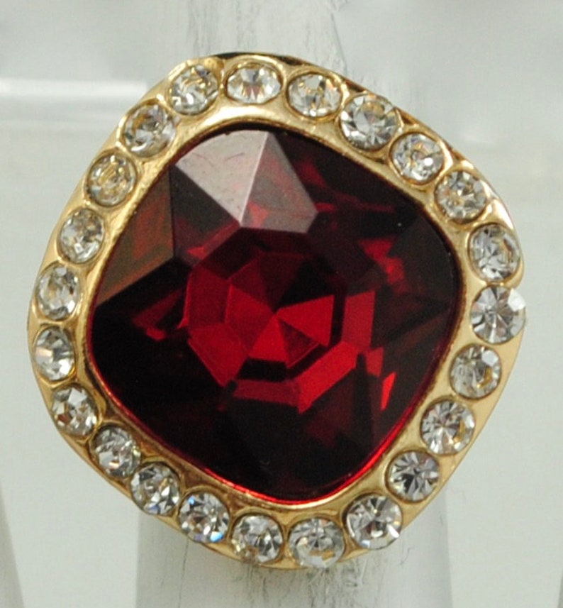 Peut inclure: Bague dor&eacute;e orn&eacute;e d'une grosse pierre rouge fonc&eacute; taill&eacute;e en carr&eacute;. La pierre est entour&eacute;e de petits cristaux transparents. Le sertissage de la bague est dor&eacute;, cr&eacute;ant un contraste avec les pierres rouges et transparentes. Un bijou de caract&egrave;re.