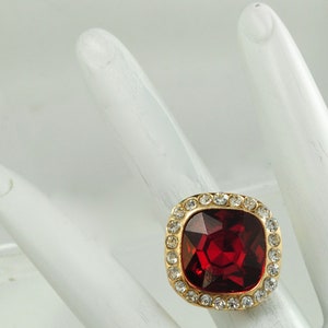 Peut inclure: Une bague dor&eacute;e orn&eacute;e d'une grande pierre pr&eacute;cieuse rouge fonc&eacute;, carr&eacute;e et facett&eacute;e. La pierre est entour&eacute;e d'un halo de petits cristaux ronds et transparents. La bague est pos&eacute;e sur une surface blanche.