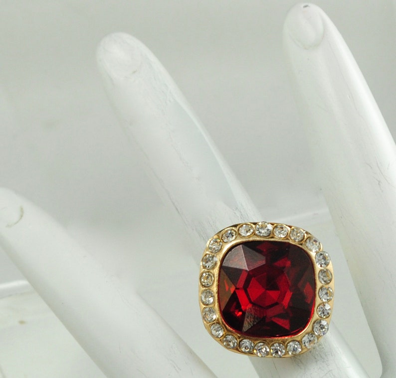 Peut inclure: Une bague dor&eacute;e orn&eacute;e d'une grande pierre pr&eacute;cieuse rouge fonc&eacute;, carr&eacute;e et facett&eacute;e. La pierre est entour&eacute;e d'un halo de petits cristaux ronds et transparents. La bague est pos&eacute;e sur une surface blanche.