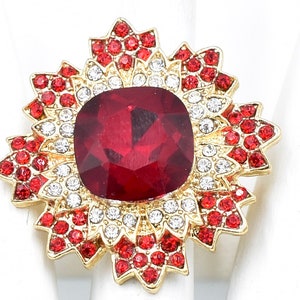 May include: A gold-toned ring featuring a large, square, deep red gemstone at its center. The gemstone is surrounded by smaller red and white crystals, arranged in a floral or starburst pattern. The ring is set against a white background.