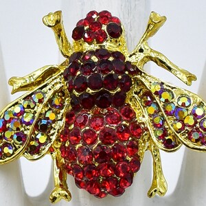 May include: A gold-toned insect-shaped ring featuring a body encrusted with deep red circular rhinestones. The wings are decorated with iridescent, multi-colored crystals that shift in hue. The metal frame is polished gold, highlighting the insect's legs.