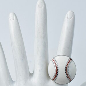 May include: A baseball-themed ring featuring a white enamel design with red stitching and silver accents. The ring is round and sits on a white hand, showcasing the details of the baseball design. The ring is a sports-themed accessory.